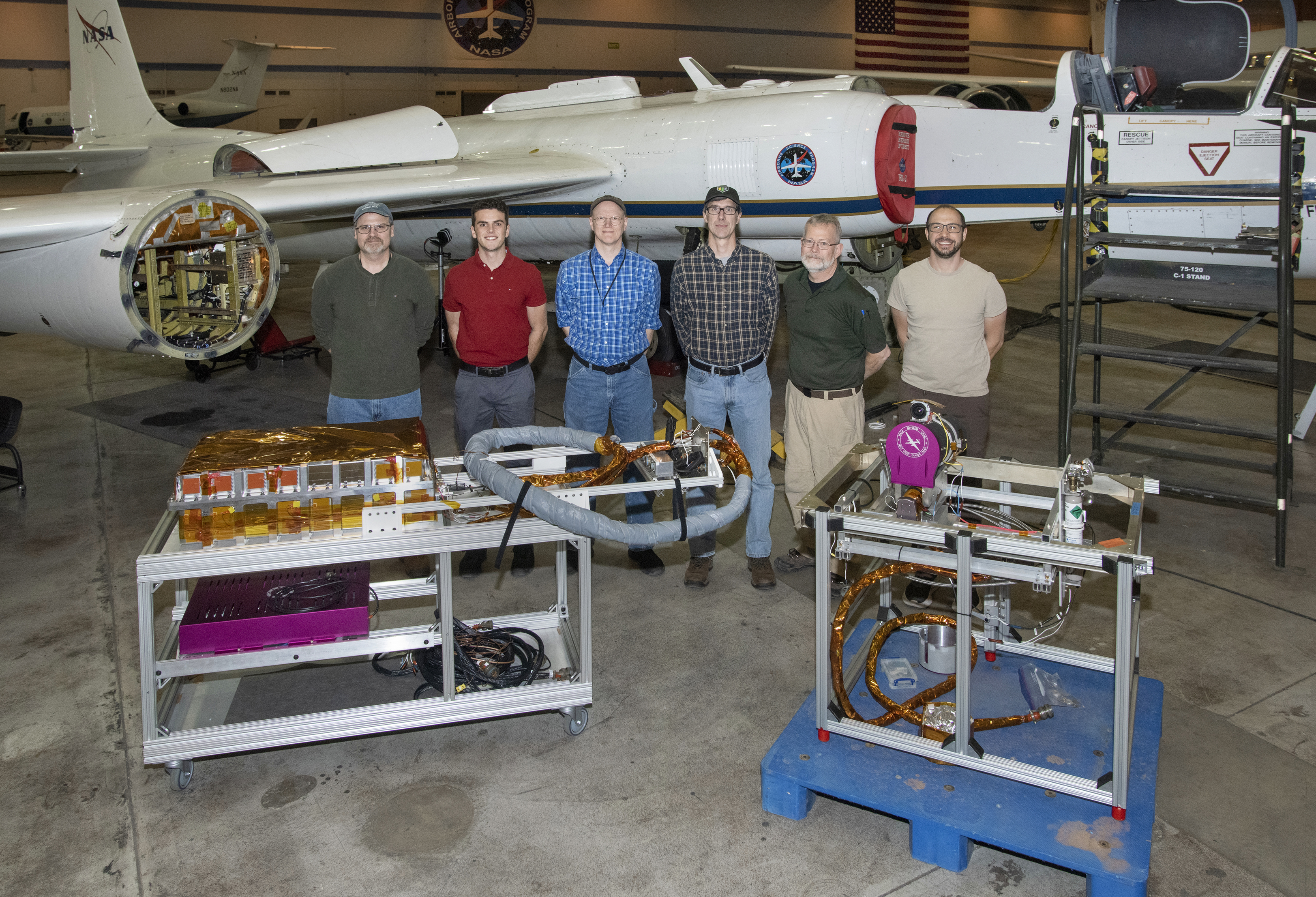 Six men stand in front of an airplane in a hangar, with equipment displayed on carts in front of them. 