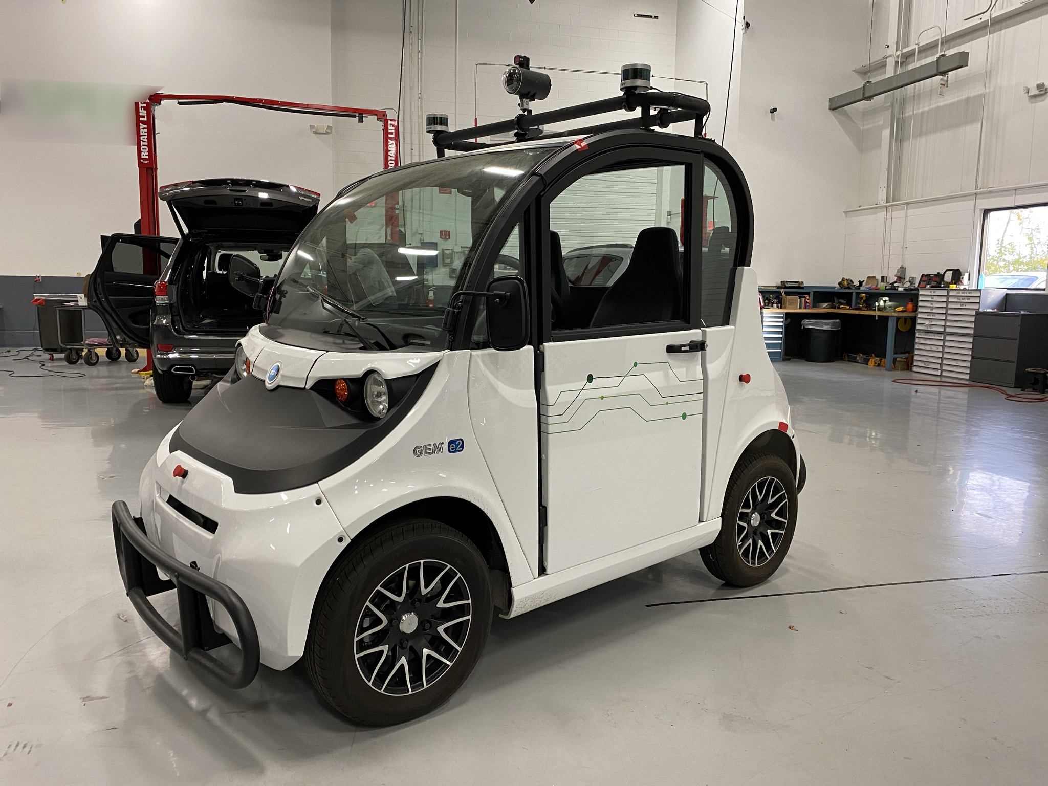 A tiny white car sits inside a large, clean garage space with tools and another vehicle in the background. 