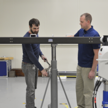 Two men stand behind a piece of metal on a custom tripod
