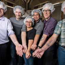 NIST researchers wearing hairnets proudly and happily present their forearms bearing tattoos of the value of the Planck constant