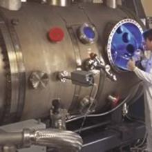 NIST physicist Charlie Tarrio stands in front of a machine that is long and silver like a sausage and has a blue light