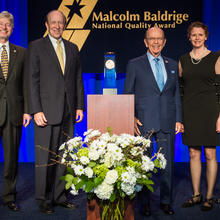 Center: Baldrige award on a podium. Left: 2 men in suits. Right: a man in a suit, woman in dress, man in suit