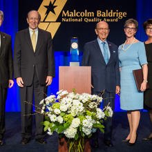 Center: Baldrige Award on a podium. Left: two men in suits. Right: a man in a suit and two women in dresses