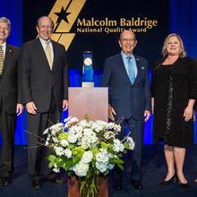 Center: Baldrige Award on a podium. Left: 2 men in suits. Right: man in suit, woman in dress, man in suit