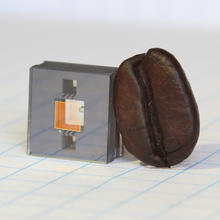 Square gray cube with two square holes in it and a gold square in between the holes, and a dark brown coffee bean leaning against the cube.