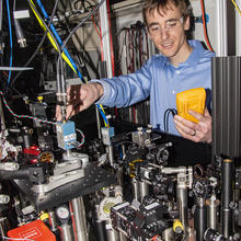 Man wearing clear goggles holding a yellow box in one hand and using the other hand to adjust a table of optics equipment