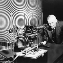 Man stands looking into the eyepiece of scientific equipment. Pattern behind is black and white concentric rings.