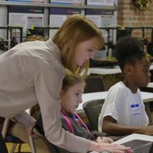 woman stands over the shoulder of two students working on laptops. 3D printers in the background.