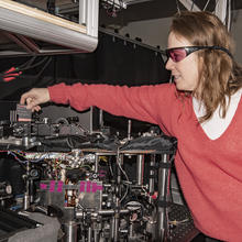 Woman with reddish hair and wearing a pink sweater and laser safety glasses reaching over an instrument table and holding a card used to path of lasers.