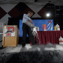 A man in safety glasses pours a foggy-looking liquid out of a metal container as part of a demonstration