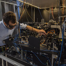 Man with dark hair, light blue shirt and laser safety glasses on the left is using his left hand to touch a device on the middle level of a three-level table covered with optics and other black and silver equipment