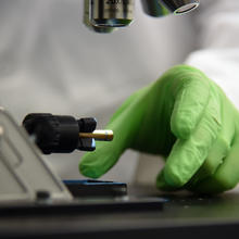 Person wearing green glove reaches under microscope to remove sample.