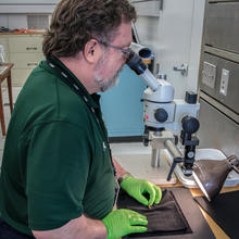 Man looks into microscope while holding bullet below.