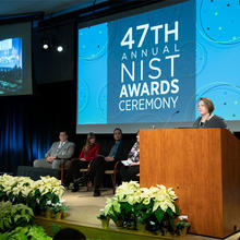 A woman stands behind a podium onstage with screen behind her saying: 47th Annual NIST Awards.