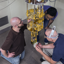 Three men work on a device that is hanging from the ceiling.