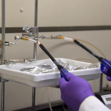 Two hands in purple gloves hold onto butane lighters, which are being used to light up two cigarettes poised in a metal device. 
