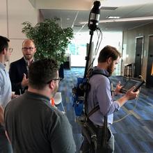 This is a photo of a LiDAR demonstration. A man is wear a backpack outfitted with LiDAR technology while three men speak around him.