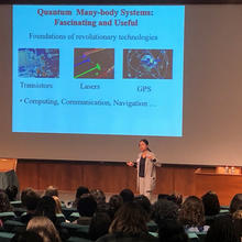 Woman on stage before a projected image that says "Quantum Many body systems: Fascinating and Useful. Foundations of revolutionary technologies: transistors, lasers, GPS. Computing, navigation, communication ...