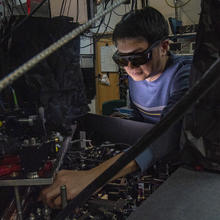 Man with dark hair and wearing a blue shirt and laser safety goggles standing on the right and facing to the left as he reaches into a two-level laboratory table full of optics and other equipment. 