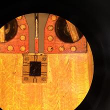 Flat, round shape in various shades of orange color, with copper wiring and other decorations covering the top third and a small, square black object surrounded by a gold frame in the center.