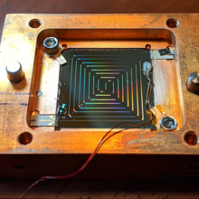 Square wood box with multicolored wires arranged in ever-smaller squares inside and three small plastic strips attached in several places.