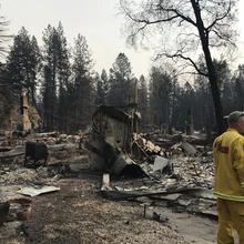 A man in a yellow fire jacket stands near burned buildings.