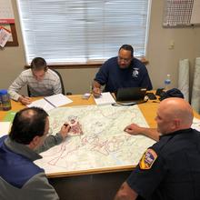 Four men sit around a table with a map spread on it.
