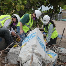Three people in hard hats and other safety gear bend over a broken concrete column marked with blue spray paint. 