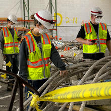 Three people in safety gear stand in a warehouse looking at chunks of concrete from Champlain Towers.