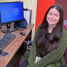 Isabelle Rivera poses smiling at a computer with two monitors, one showing a spreadsheet and the other multiple file icons. 