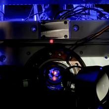 Close-up of a scientific instrument with wires and a circular metallic component, illuminated by blue light, featuring a small red dot at the center.