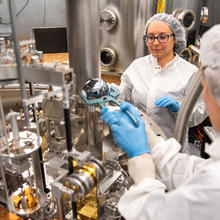 A researcher wearing a lab coat uses tongs to place a silvery sphere into a large scientific device as another researcher looks on. 