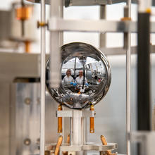 The faces of two researchers in lab coats are reflected in a silvery sphere that is being held inside a larger scientific device. 
