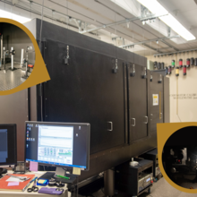 A high-tech laboratory with large black equipment featuring two circular insets showing detailed machinery setups. A computer workstation is in the foreground.
