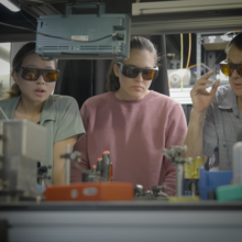 Three researchers wearing safety glasses stand in a row looking down at a complex setup of lab equipment. 