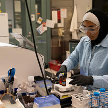 SURF student Zainab Altamimi, who wears a hijab, stands in a lab with safety glasses and gloves on as she holds a micropipette in one hand and a vial in the other.
