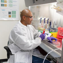 A researcher wearing safety gear sits at a lab table working with a tray of samples and a large vial holding red liquid.