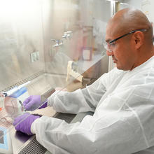 A researcher wearing safety gear works with small tools in the lab, putting his hands inside a plexiglass box. 
