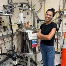 Cassie Stoffer smiles while wearing safety glasses and standing next to a large metal tank with many pipes and wires attached to it. 