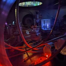 Table in darkened lab holds wiring and other laser electronics; a circular light source is situated above. 