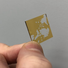 A tiny flat square with a chipped coating of gold is held between a finger and thumb. 