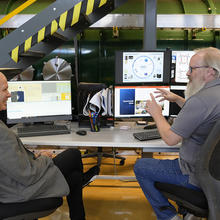 Two men sit at a table in a lab, facing each other and smiling as they talk and gesture toward their computer screens. 