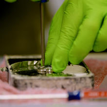 Close-up shows a hand in a green glove holding the center of a metal disk while using a pointed tool to remove a part. 