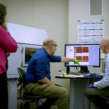 A man points to images on a computer screen in a lab as two other researchers look on, one seated and one standing. 