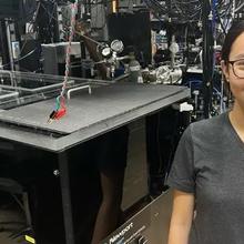 A woman poses in a lab full of electronics and wiring, making a thumbs-up gesture. 