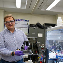 Peter Beaucage wears safety glasses as he stands in the lab, holding a piece of equipment on display. 