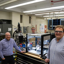 Three men in safety glasses pose standing around a large clear plastic box on a lab table. The box holds several small pieces of equipment. 