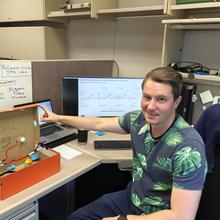 Sam Schaffter poses sitting in his office with a shoebox modified with craft supplies to be an educational model of an E. coli cell. 