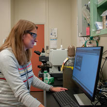 A scientist wearing safety glasses stands in front of a monitor in her lab.