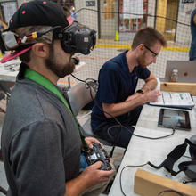 A man wearing an electronic device over his eyes sits at a table holding a controller for a drone. The indoor training facility includes pretend storefronts like a pizza shop. 